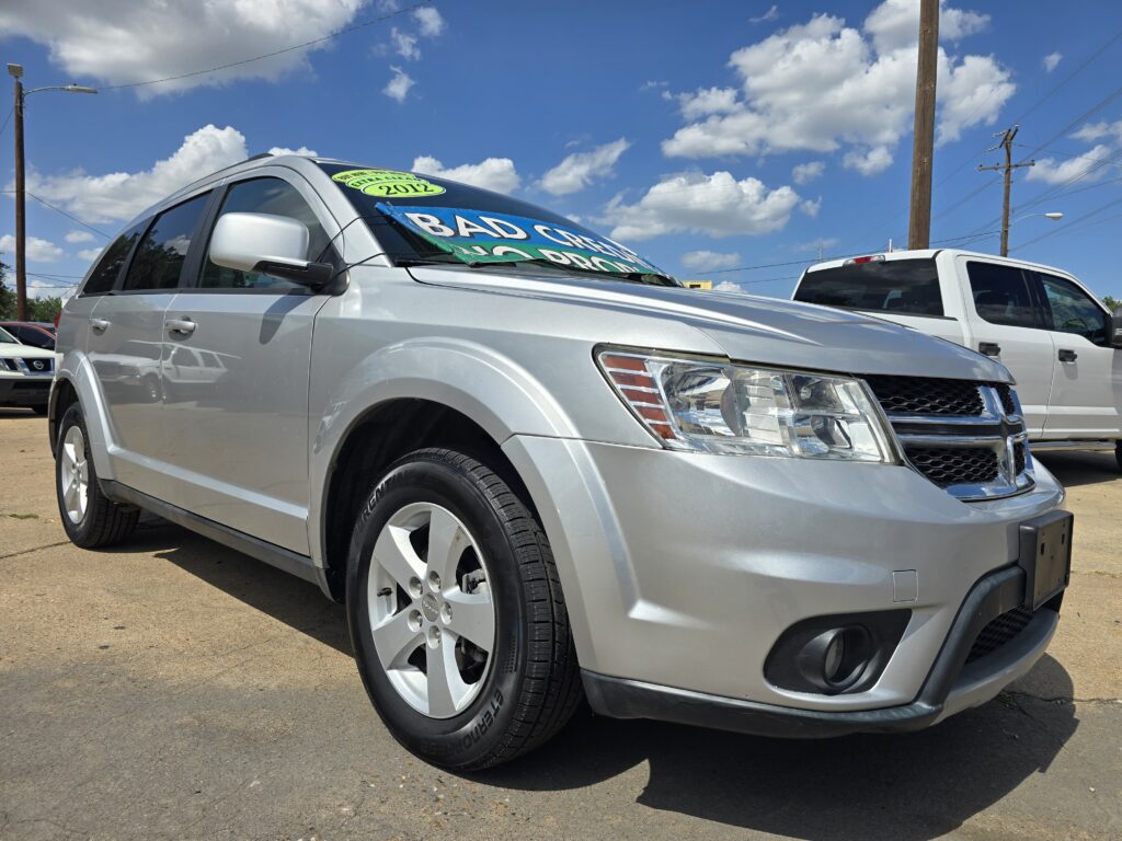 Front-left view of a silver SUV with a green/yellow circular '2012' sticker on the windshield, at a sunny car lot under a blue sky.