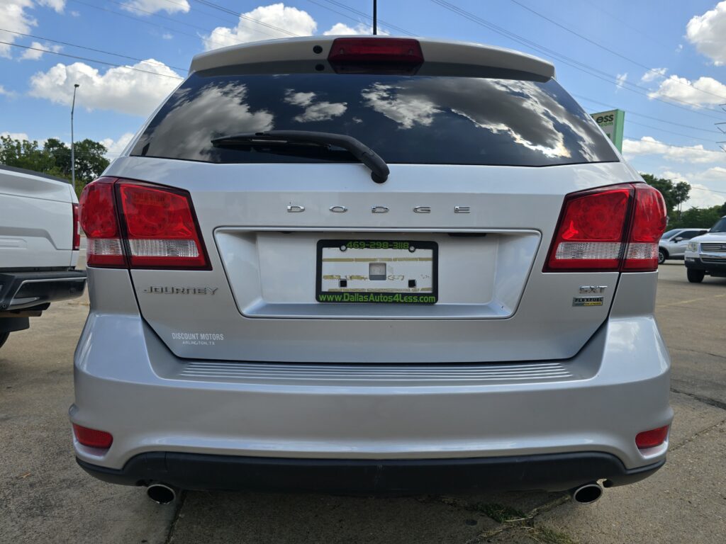 Rear view of a silver Dodge Journey SUV with red taillights, rear window wiper, and dealer plate frame that reads Dallas Autos 4 Less (Arlington, TX).