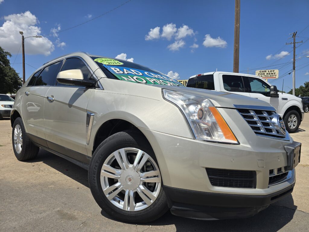 Silver Cadillac SUV in a dealership lot under a blue sky; windshield shows a neon green sale sign reading 2014, with another white vehicle nearby.