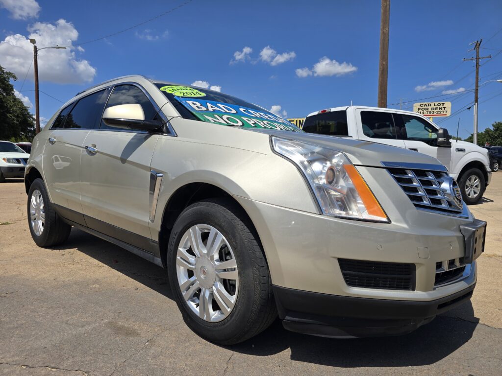 Silver Cadillac SUV parked at a car dealership lot on a sunny day, with a large windshield sign visible in the front window.