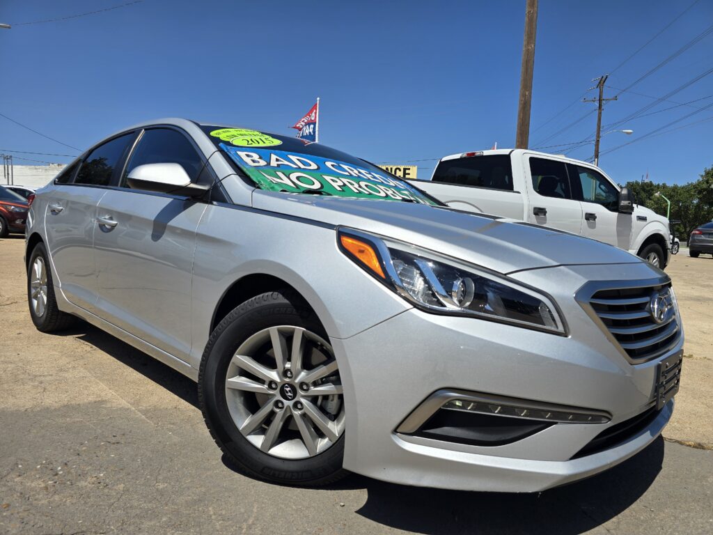 Silver sedan with a bold green-and-blue windshield banner reading 'BAD CREDIT NO PROBLEM' at a car lot under a clear blue sky, with a white pickup beside it.