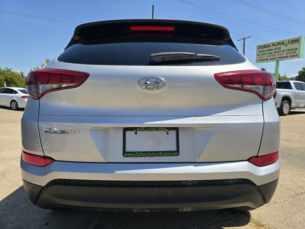 Rear view of a silver Hyundai Tucson in a car lot, showing the Hyundai logo and taillights; dealership sign in the background reads Dallas Autos 4 Less.