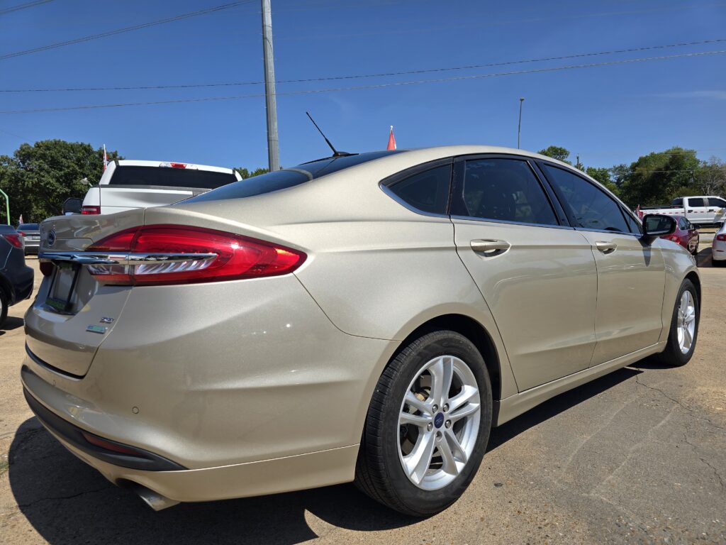 Beige Ford sedan viewed from rear side in a sunny parking lot with other cars nearby and a clear blue sky.