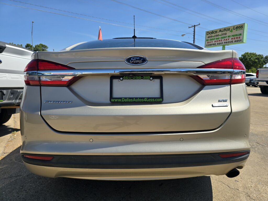 Rear view of a silver Ford Fusion at a car lot, with a green Dallas Autos 4 Less license plate frame and dealership sign in the background